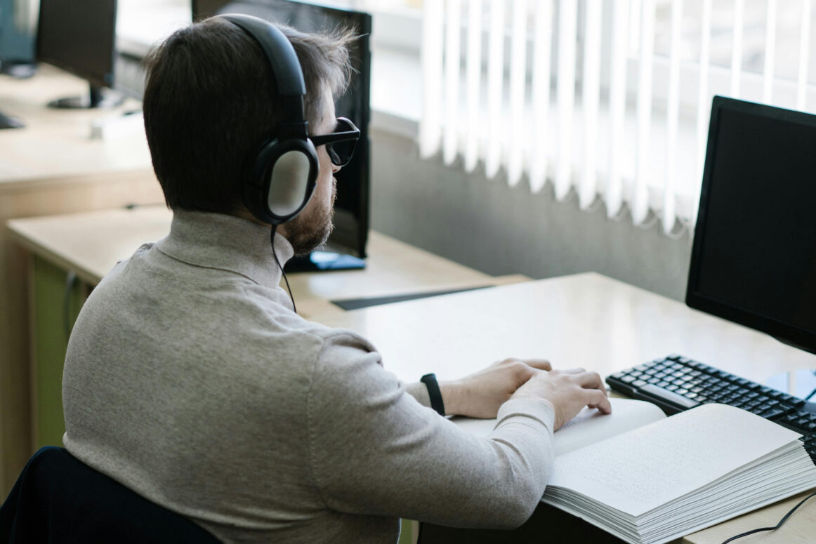 Blind mand sidder ved et bord og læser braille med headset på.
